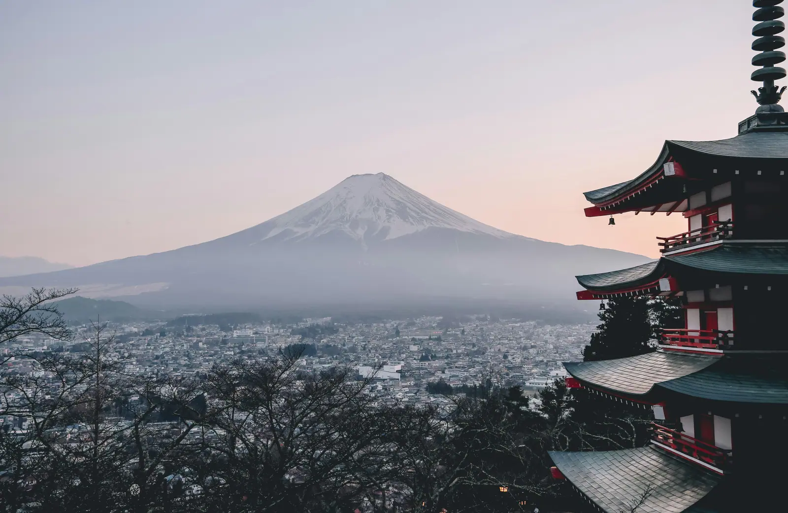 Mount Fuji before sunrise above Fujiyoshida, seen past the tiered roofs of Chureito Pagoda. The mountain reads as flat planes of grey haze over the town below.