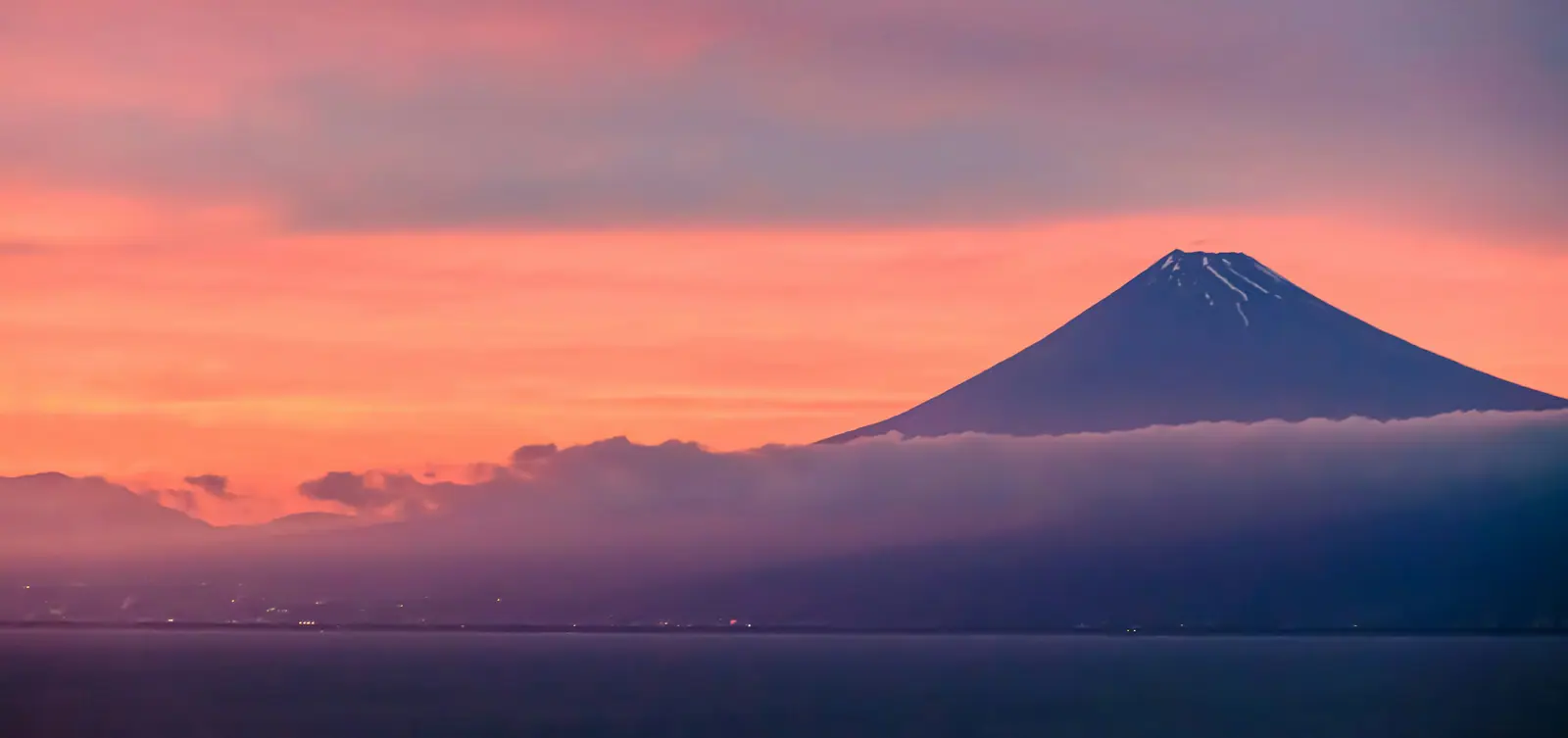 Mount Fuji across the water at dawn under a wide pink and apricot sky, a bar of cloud lying along the base of the cone.