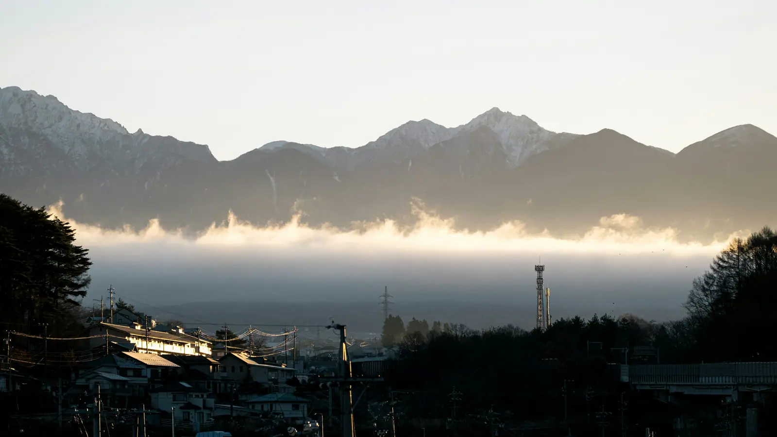 A snow-topped mountain range above a long bar of low cloud lit gold from beneath, with the roofs and power lines of a small town in the foreground.