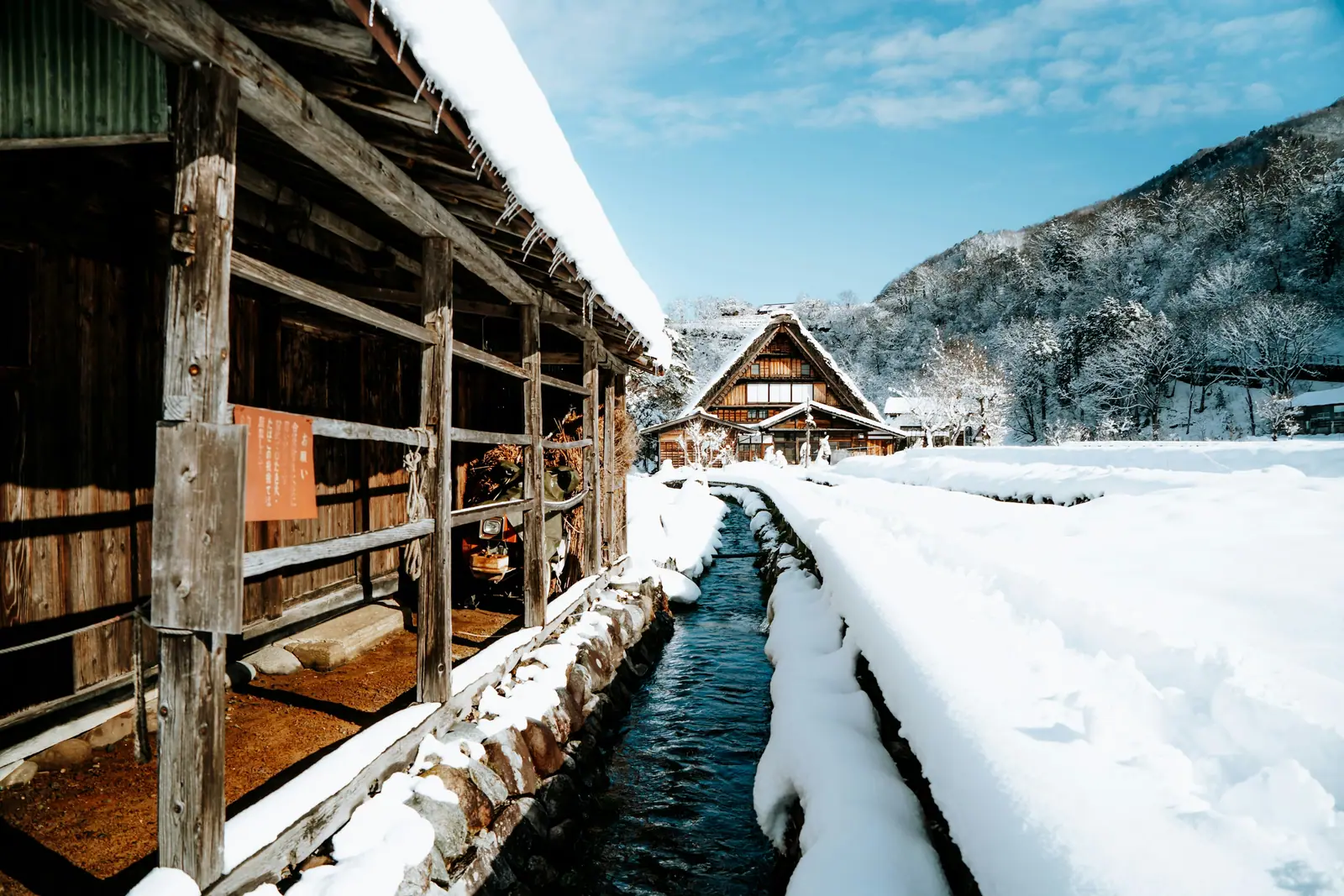 A meltwater channel running between deep snowbanks through Shirakawa-gō, past a timber storehouse, towards a thatched gasshō-zukuri farmhouse under a wooded slope.
