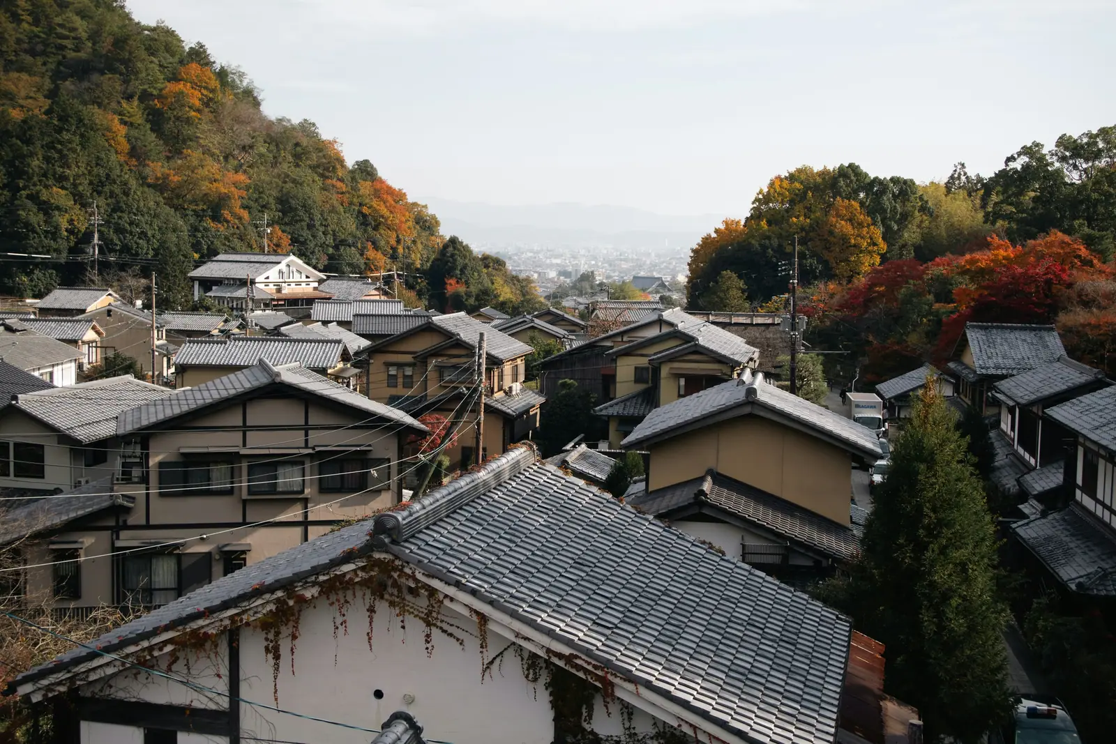 Tiled rooftops of houses stepping down a wooded Kyoto hillside in autumn, with the city and distant mountains showing pale through the haze beyond.