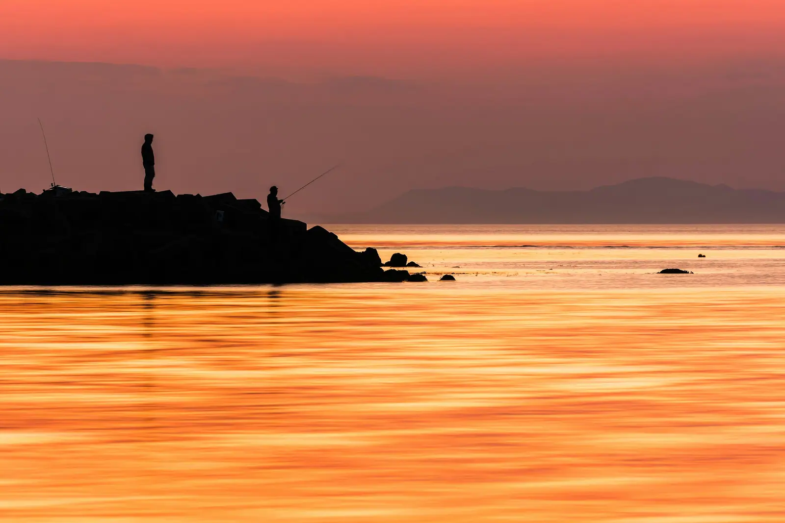 Two anglers silhouetted on a rock breakwater at sunset, the water in front of them a sheet of orange light and a headland fading into haze on the horizon.