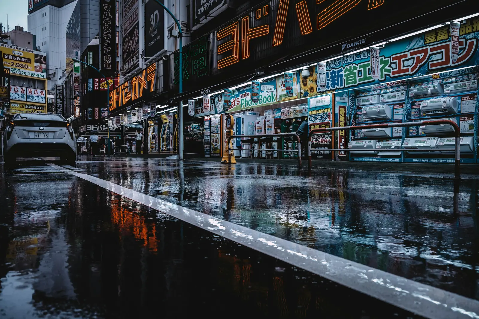A rain-soaked Tokyo street at night, neon shopfront signage reflected in long streaks across the wet asphalt.