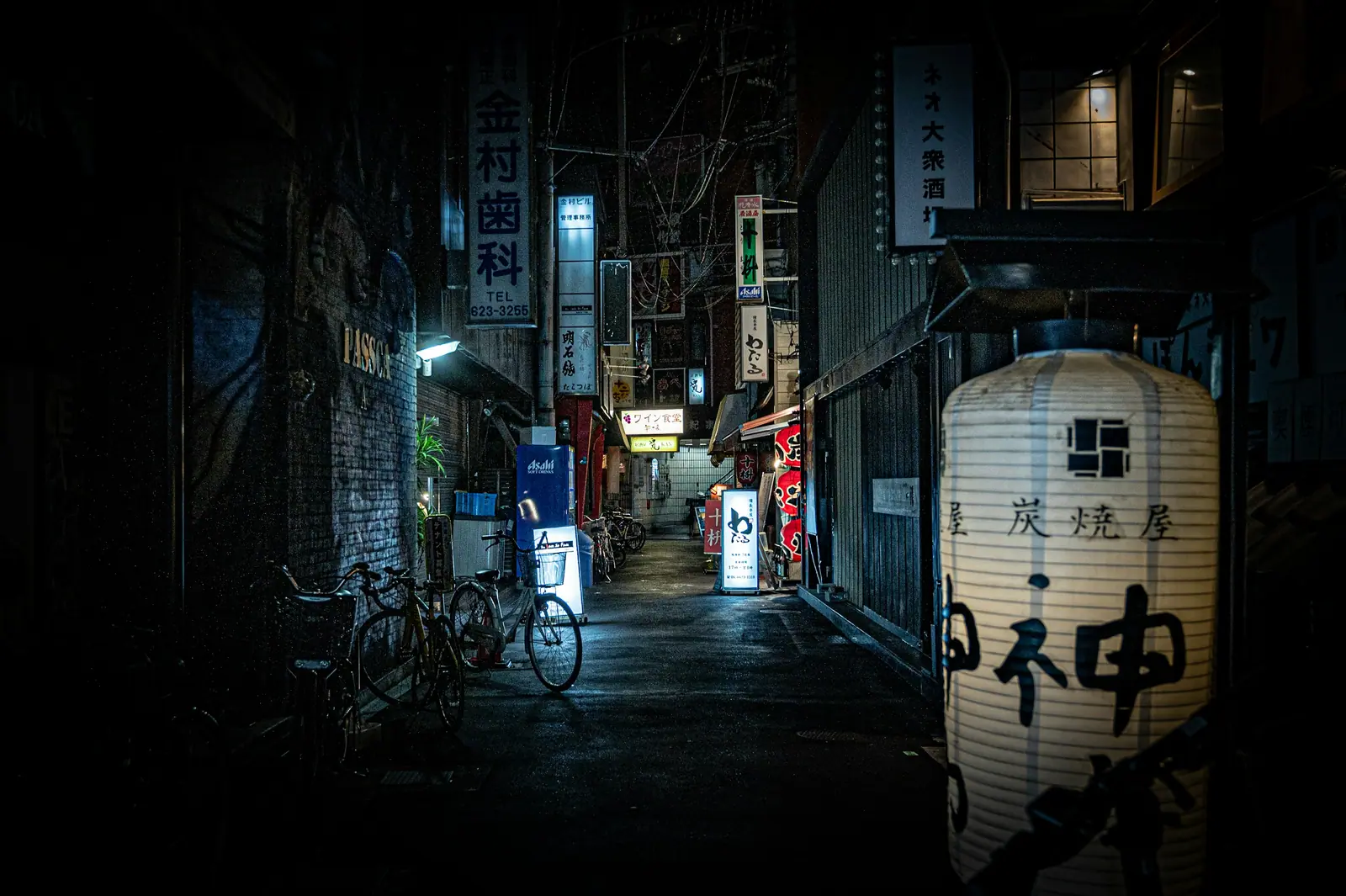 A narrow Osaka alley at night, bicycles against a brick wall on the left and a tall paper lantern glowing at the right, shop signs receding into the dark.